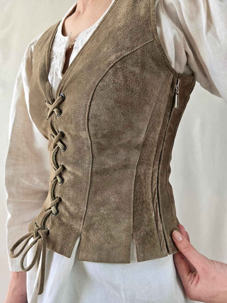 May include: A sleeveless olive brown suede vest featuring a front lace-up closure with matching suede ties, metal grommets, and a side zipper. The garment is constructed with vertical paneling and a straight hemline with small side slits.