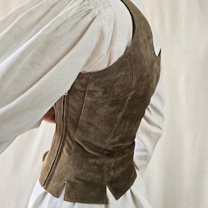 May include: A dark olive green suede vest featuring a fitted silhouette with vertical seam detailing, a side zipper closure, and a straight hemline with small slits, worn over a white long-sleeved shirt with gathered shoulders.