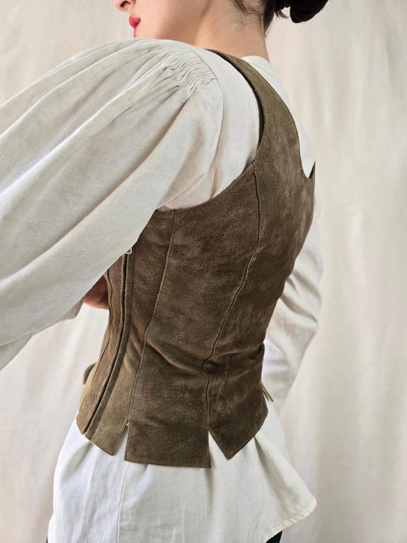 May include: A dark olive green suede vest featuring a fitted silhouette with vertical seam detailing, a side zipper closure, and a straight hemline with small slits, worn over a white long-sleeved shirt with gathered shoulders.