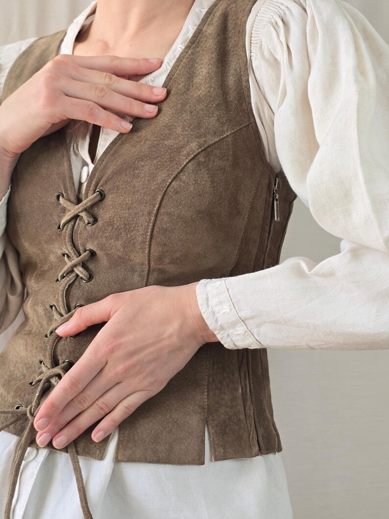 May include: A brown suede vest featuring a front lace-up closure with metal eyelets and a side zipper. The vest is worn over a white long-sleeved shirt with gathered shoulders and cuffs. The vest has a V-neckline and a straight hem with side slits.
