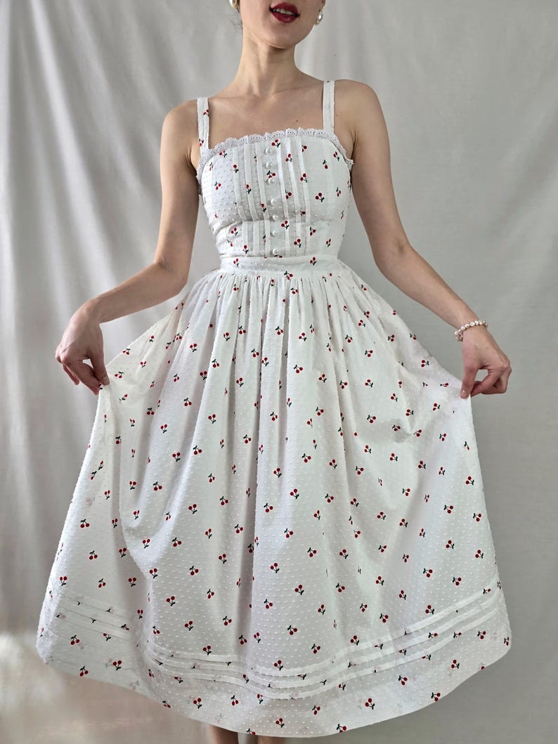 May include: A white dress with a cherry print and a fitted bodice. The dress has a full skirt and a lace trim at the neckline and hem.
