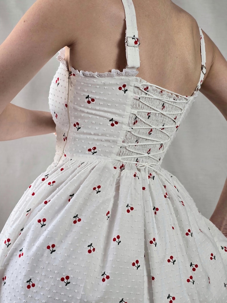 May include: White dress with a cherry print and a lace trim. The dress has a fitted bodice with a lace-up back and a full skirt.