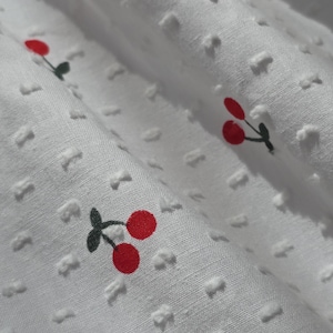 May include: White fabric with a textured pattern and a repeating pattern of red cherries with green stems.