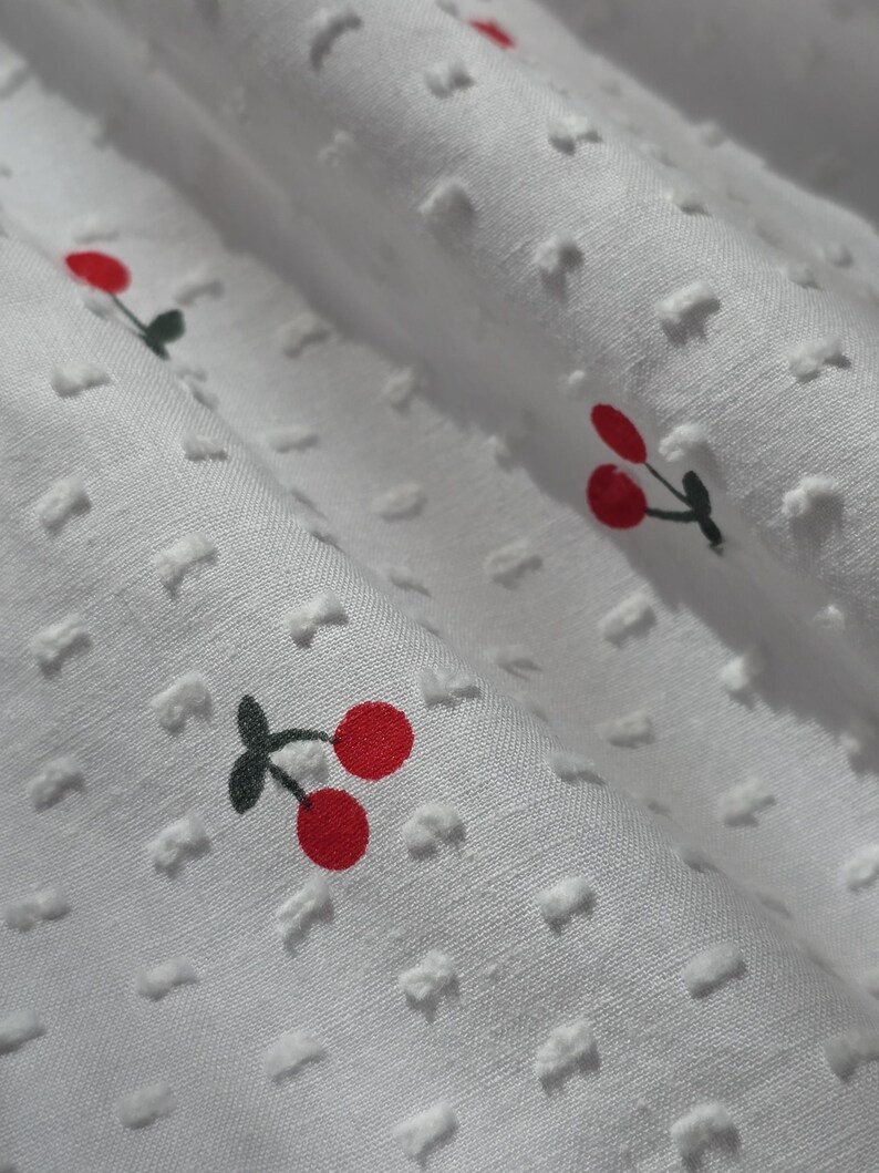 May include: White fabric with a textured pattern and a repeating pattern of red cherries with green stems.