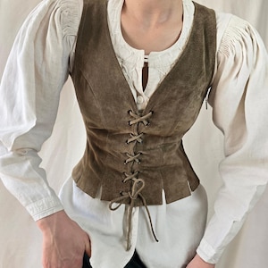 May include: A brown suede vest featuring a front lace-up closure with matching brown cord ties, worn over a white long-sleeved blouse with gathered shoulders and a button-down front, paired with a dark green skirt.