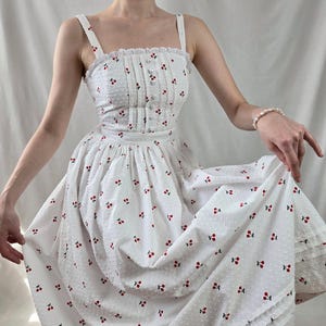 May include: A white midi dress with a cherry print and a lace trim. The dress has a fitted bodice and a full skirt.