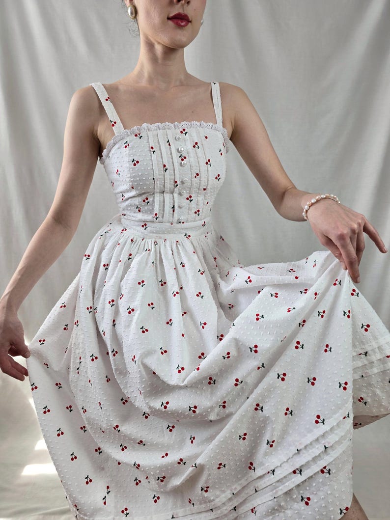 May include: A white midi dress with a cherry print and a lace trim. The dress has a fitted bodice and a full skirt.