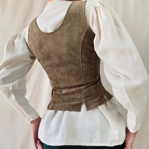 May include: A sleeveless brown suede vest featuring a scoop neckline and a fitted bodice with small side slits at the hem. The vest is worn over a white long-sleeved shirt with puffed sleeves and is paired with a dark green skirt.