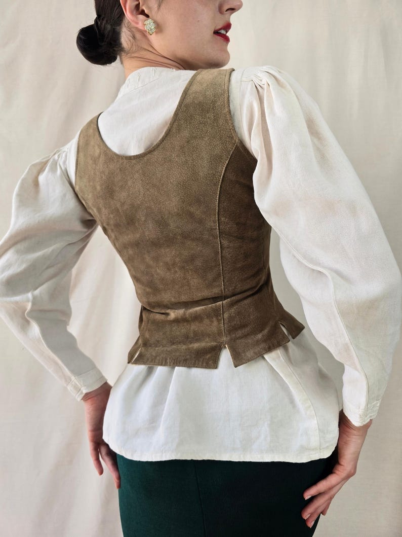 May include: A sleeveless brown suede vest featuring a scoop neckline and a fitted bodice with small side slits at the hem. The vest is worn over a white long-sleeved shirt with puffed sleeves and is paired with a dark green skirt.