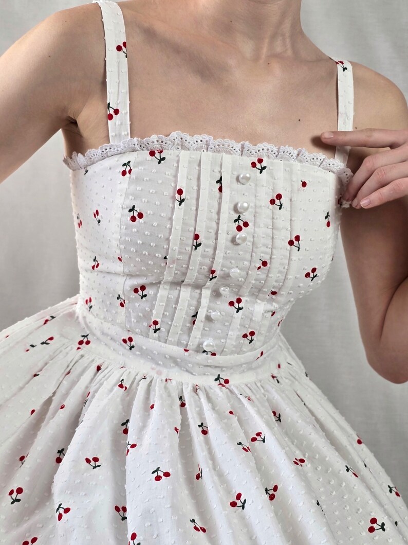 May include: A white sundress with a cherry print and a lace trim. The dress has a fitted bodice and a full skirt.