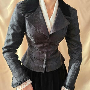 May include: A dark gray floral patterned jacket with a black velvet collar and ruffled cuffs. The jacket has a fitted waist and is paired with a black skirt and a white blouse.
