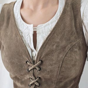 May include: A brown suede vest featuring a front lace-up closure with dark metal eyelets and a V-neckline. The vest is worn over a white blouse with a round neckline, vertical pleats, and white decorative stitching along the collar edge.