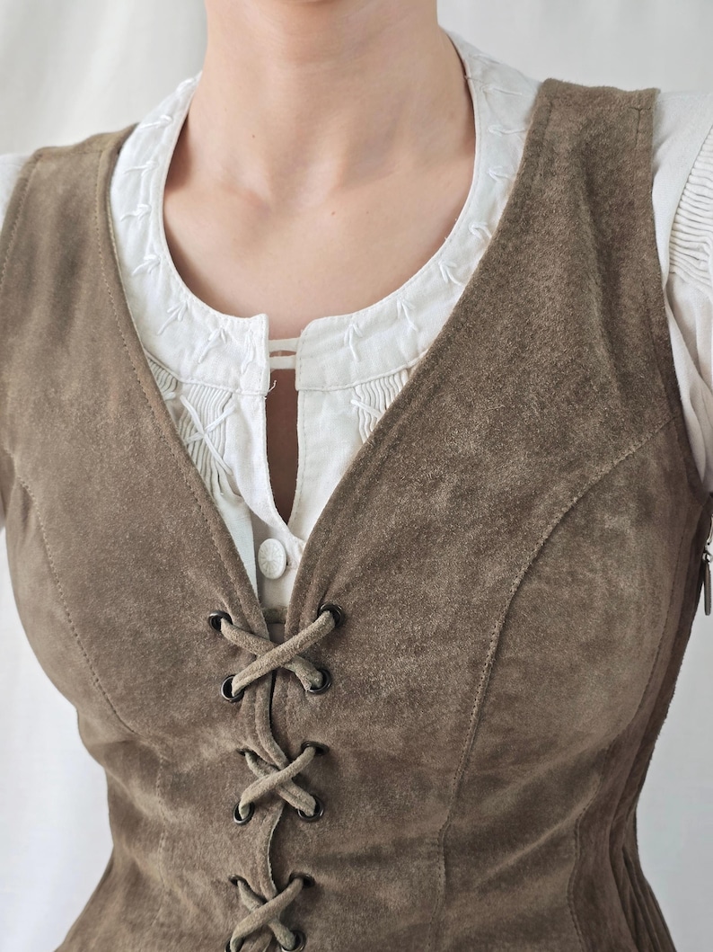 May include: A brown suede vest featuring a front lace-up closure with dark metal eyelets and a V-neckline. The vest is worn over a white blouse with a round neckline, vertical pleats, and white decorative stitching along the collar edge.