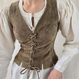 May include: A brown suede vest featuring a front lace-up closure with matching suede cord ties, a deep V-neckline, and a hem with vertical slits. The vest is worn over a white long-sleeved shirt with gathered shoulders and a button-down front.