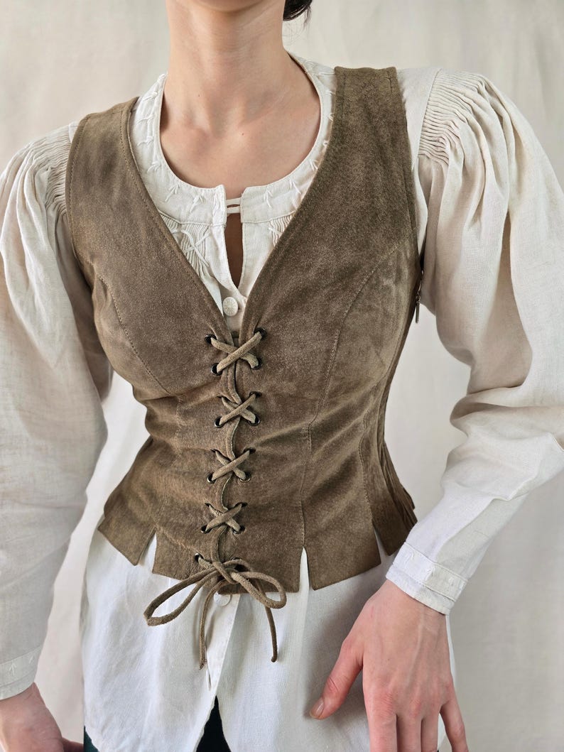 May include: A brown suede vest featuring a front lace-up closure with matching suede cord ties, a deep V-neckline, and a hem with vertical slits. The vest is worn over a white long-sleeved shirt with gathered shoulders and a button-down front.