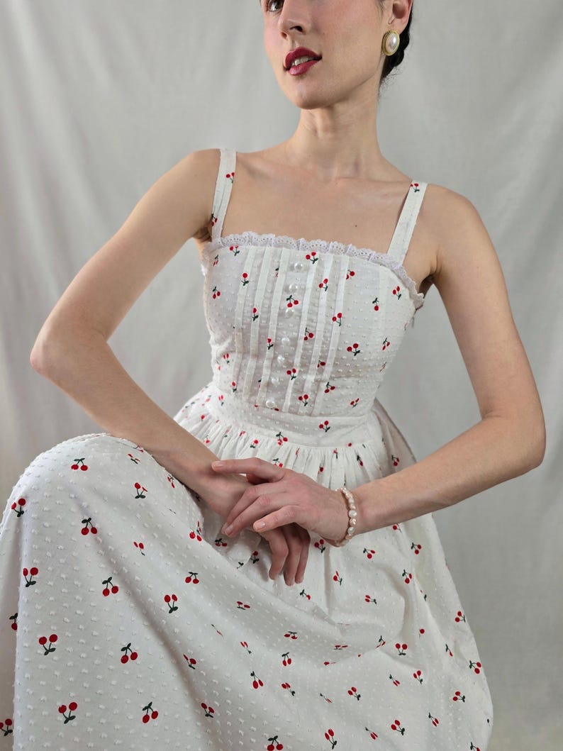 May include: A white midi dress with a cherry print and a lace trim. The dress has a fitted bodice and a full skirt.