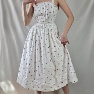 May include: A white midi dress with a cherry print and a lace trim. The dress has a fitted bodice and a full skirt.