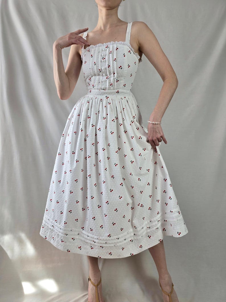 May include: A white midi dress with a cherry print and a lace trim. The dress has a fitted bodice and a full skirt.