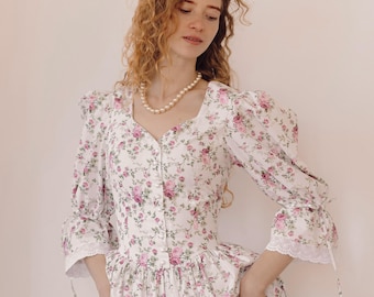 May include: A white floral print dress with pink roses. The dress features a square neckline, puff sleeves, and a button-down front. The sleeves and hem are trimmed with lace. A pearl necklace is worn.