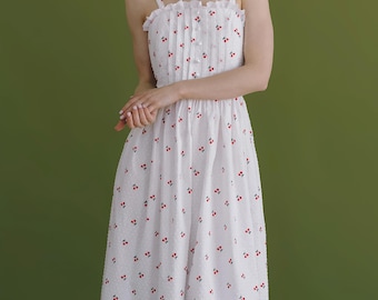May include: White sundress with red cherry print. The dress features a square neckline, thin straps, and a button-down front. The dress has a midi length and a ruffled trim at the top. The model is wearing a pearl necklace.