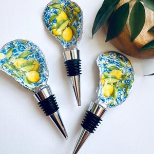 May include: Three bottle stoppers with a lemon design. Each has a silver base, a black rubber seal, and a top with a blue and white pattern featuring yellow lemons and green leaves.