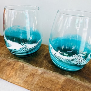 May include: Two clear glass wine glasses with a teal blue and white ocean wave design. The glasses are sitting on a wooden surface.