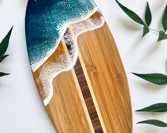 May include: An oval wooden serving board with a resin ocean design. The top section has a deep blue resin, representing the sea, which flows into a sandy-coloured resin with white wave details. The board is made of bamboo wood, with a central dark brown stripe.