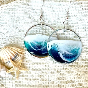 May include: A pair of silver hoop earrings with a blue and white resin design that resembles ocean waves. The resin is clear and has a textured surface.