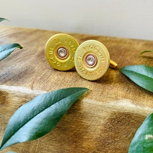 Peut inclure: Une paire de boutons de manchette dorés fabriqués à partir d'étuis de cartouches de fusil de chasse. Les boutons de manchette ont un design circulaire avec le texte "FIOCCHI ITALY 12" imprimé dessus. Les boutons de manchette sont présentés sur une surface en bois.