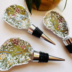 May include: Three bottle stoppers with a floral pattern in green, red, and yellow. Each stopper has a silver metal base and a black rubber seal. The stoppers are designed for wine bottles. The background is white.