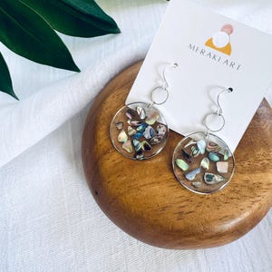 May include: A pair of round, silver-toned earrings. Each earring features a clear, circular resin design filled with iridescent shell fragments in shades of blue, green, pink, and brown. The earrings are displayed on a wooden surface with a white card that reads "MERAKI ART".