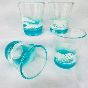 May include: Four clear glass tumblers with a blue and white wave design. The glasses are on a white fabric surface. Ocean Scene Shot Glass Barware Beach House Decor Kitchen Decor Coastal Decor Gifts For Her Gifts For Him Beach Lovers Glassware