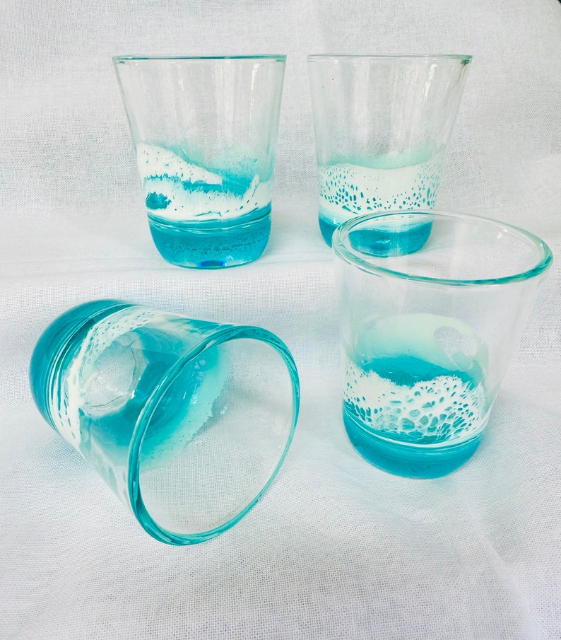 May include: Four clear glass tumblers with a blue and white wave design. The glasses are on a white fabric surface. Ocean Scene Shot Glass Barware Beach House Decor Kitchen Decor Coastal Decor Gifts For Her Gifts For Him Beach Lovers Glassware