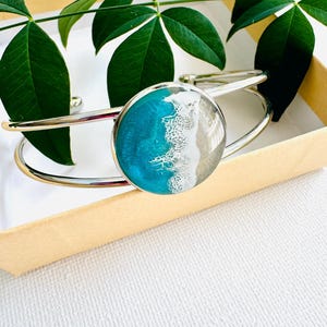 May include: A silver bangle bracelet with a round, turquoise and white resin cabochon. The cabochon features a wave design.