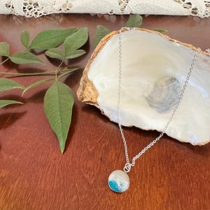 May include: A silver chain necklace with a round pendant. The pendant features a design with shades of blue and white, resembling ocean waves. The necklace is displayed on a wooden surface with a decorative seashell.