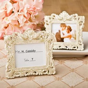 May include: Two ornate white picture frames with a vintage design. The frames are made of plastic and have a raised, detailed border. The frames are perfect for displaying photos or small artwork. The frame on the left has a blank card with the text "Mr. H. Cassidy" and "Table 7" written on it. The frame on the right has a photo of a couple.