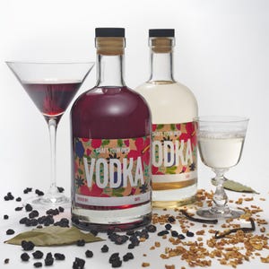 May include: Two glass bottles of vodka with colourful floral labels that say "Craft Your Own Vodka". One bottle is clear and the other is a deep purple colour. The bottles are sitting on a white surface with dried fruit and berries scattered around them.