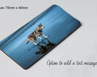 Op de afbeelding: Een grijze en witte wolfshond staat in een mistig blauw meer. De hond kijkt naar rechts in de afbeelding. De reflectie van de hond is zichtbaar in het water. De afbeelding is een magneet met een afmeting van 75 mm x 40 mm.