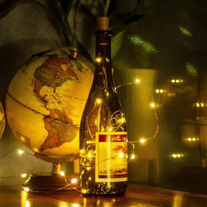 May include: A brown wine bottle with a cork stopper, illuminated by fairy lights. The bottle has a label with text and is next to a globe. The lights are glowing, creating a warm ambiance.