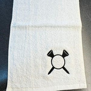 Peut inclure: Serviette de golf blanche avec un motif brodé noir de deux tees de golf croisés sur une balle de golf.