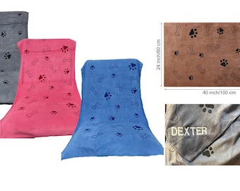 May include: Three dog towels in gray, pink, and blue with paw print and bone designs. The towels are 40 inches by 60 inches. A brown towel with paw print and bone designs is shown in the background. The towels are personalized with the names "Dexter" and "Frank".