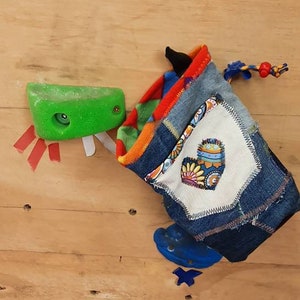 May include: A colorful, patchwork climbing chalk bag with a heart-shaped design and a blue drawstring. The bag is made from denim and other fabrics. It is hanging on a wooden wall.