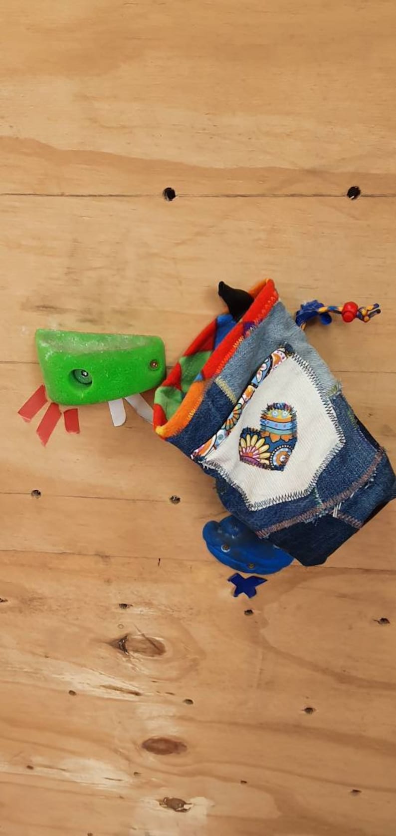 May include: A colorful, patchwork climbing chalk bag with a heart-shaped design and a blue drawstring. The bag is made from denim and other fabrics. It is hanging on a wooden wall.