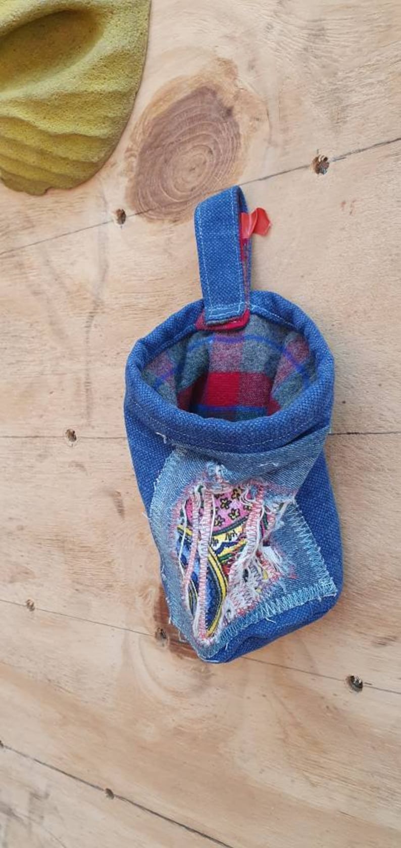 May include: A blue denim pouch with a red and black plaid lining. The pouch has a loop handle and a patch of colorful fabric sewn on the front.