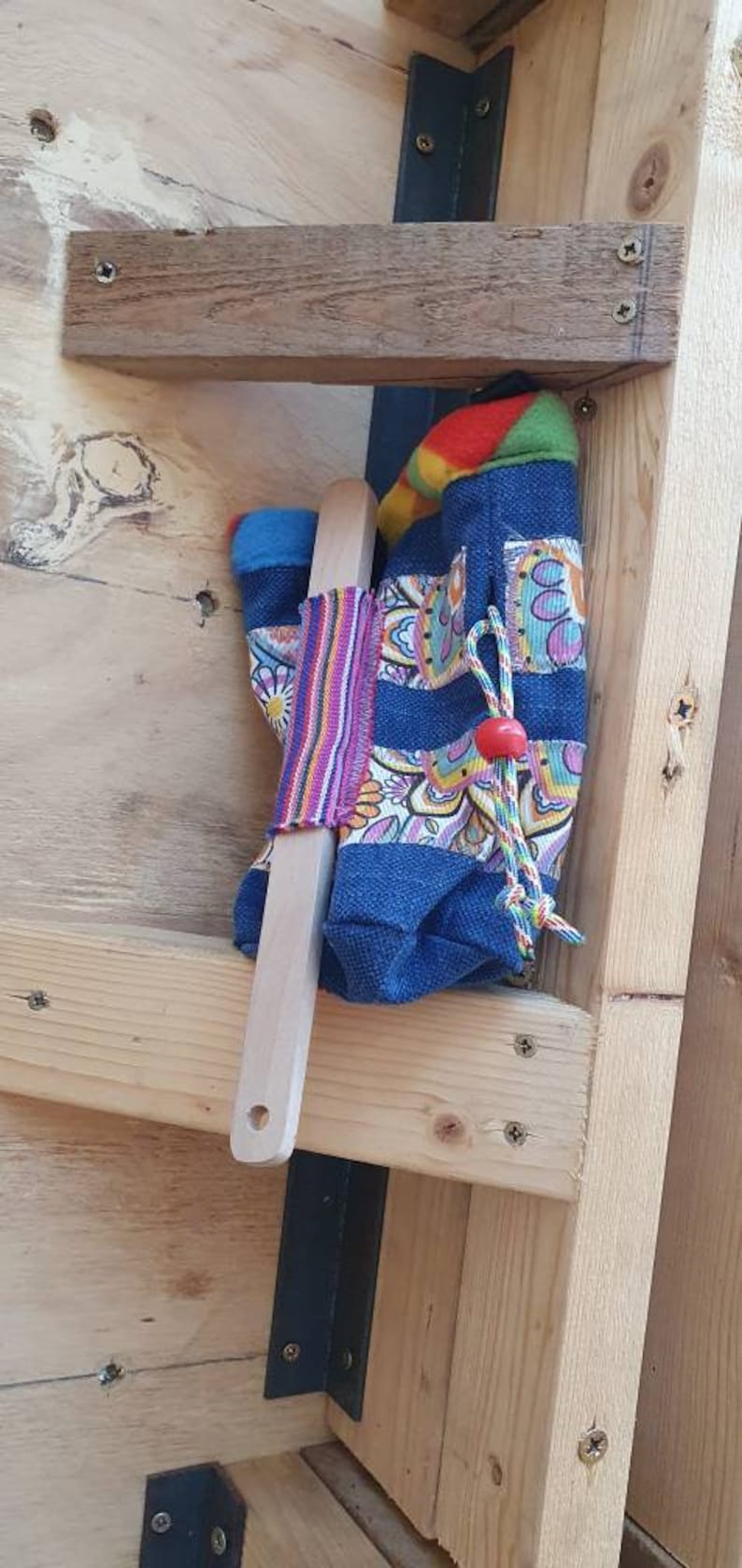 May include: A wooden shelf with a blue and colorful fabric bag hanging from it. The bag has a wooden dowel inside and a red bead on the drawstring. The shelf is made of wood and has a metal bracket attached to it.