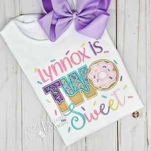 May include: A white short-sleeved shirt featuring embroidered text that reads "Lynnox is TWO Sweet" with a donut graphic. The shirt is decorated with colorful sprinkles and includes a large, solid light purple ribbon hair bow attached at the top.
