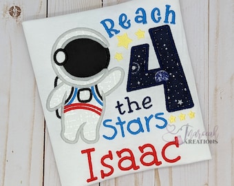 May include: A white t-shirt featuring an embroidered astronaut, a large dark blue number 4 with a space pattern, and the text Reach the Stars Isaac. The astronaut is white with black, red, and blue details. Yellow stars accent the design.