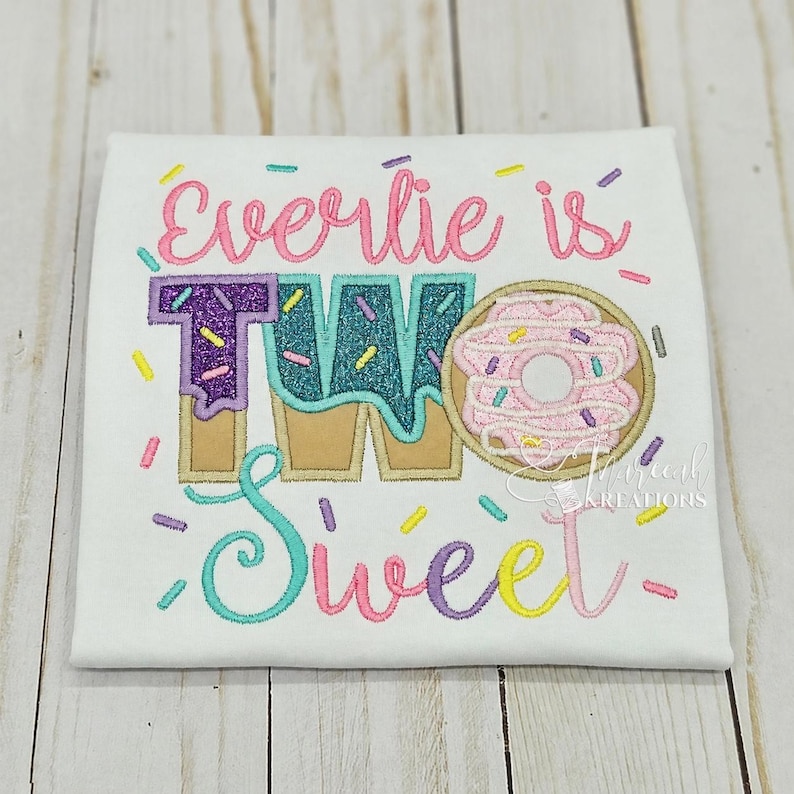 May include: A white t-shirt featuring embroidered text that reads Everlie is TWO Sweet. The design includes a large donut graphic, glittery teal and purple lettering, and colorful sprinkles scattered across the fabric in pink, yellow, and teal.