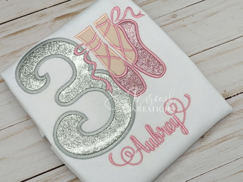 Puede incluir: Una camiseta blanca con un n&uacute;mero "3" de purpurina plateada y zapatillas de ballet rosas con cintas rosas. El nombre "Aubrey" est&aacute; bordado en rosa debajo del n&uacute;mero.