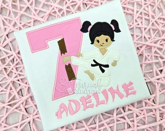 May include: A white t-shirt featuring an embroidered design of a girl in a white karate uniform with a black belt, performing a kick that breaks a brown wooden board. A large pink number seven and the name ADELINE in pink letters are embroidered.